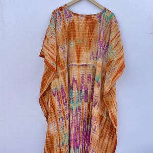 Puede incluir: Un caft&aacute;n te&ntilde;ido anudado en tonos naranja, morado y turquesa cuelga de una percha de madera. La tela presenta un patr&oacute;n vertical con un dise&ntilde;o similar a una cuadr&iacute;cula. La prenda tiene mangas anchas y una silueta suelta y fluida, adecuada para ropa informal.