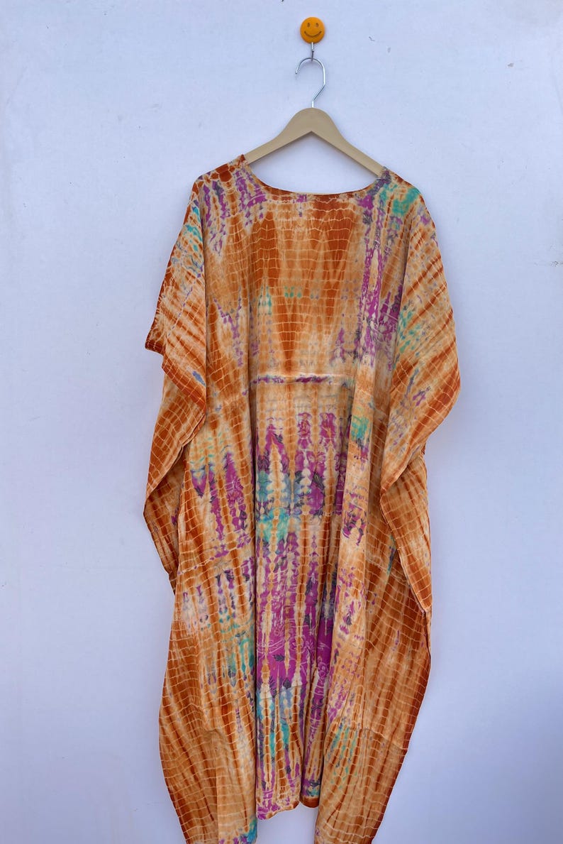Puede incluir: Un caft&aacute;n te&ntilde;ido anudado en tonos naranja, morado y turquesa cuelga de una percha de madera. La tela presenta un patr&oacute;n vertical con un dise&ntilde;o similar a una cuadr&iacute;cula. La prenda tiene mangas anchas y una silueta suelta y fluida, adecuada para ropa informal.