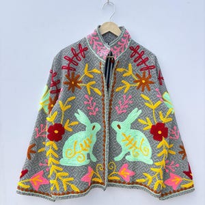 Può includere: Giacca grigia con ricami colorati raffiguranti due conigli verdi, fiori e foglie. La giacca ha un collo alto ed è appesa a una gruccia di legno. Il ricamo include dettagli rosa, gialli, rossi e marroni.