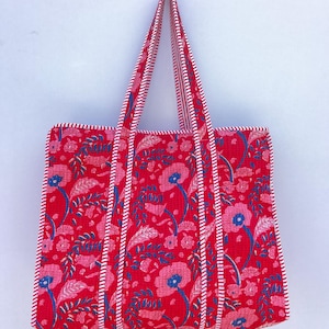 May include: A red tote bag with a floral pattern featuring pink and blue flowers and leaves. The bag has white and red striped handles and trim. The bag is hanging against a white background.