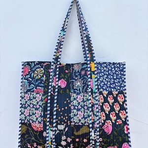May include: A quilted tote bag featuring a patchwork design of various floral patterns in shades of navy blue, pink, white, and green. The bag has two long shoulder straps with a blue and white striped pattern and is hung from a yellow smiley face hook.
