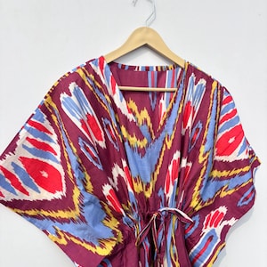 May include: A burgundy kaftan with a V-neck and a drawstring waist. The fabric features a vibrant pattern of red, blue, yellow, and white abstract shapes. The garment is hanging on a wooden hanger against a white background.