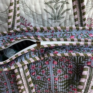 May include: A quilted tote bag with a floral pattern in pink and green on a light blue background. The bag has a white zipper and olive green straps with pink and white stripes. The bag is made of a soft, quilted material.