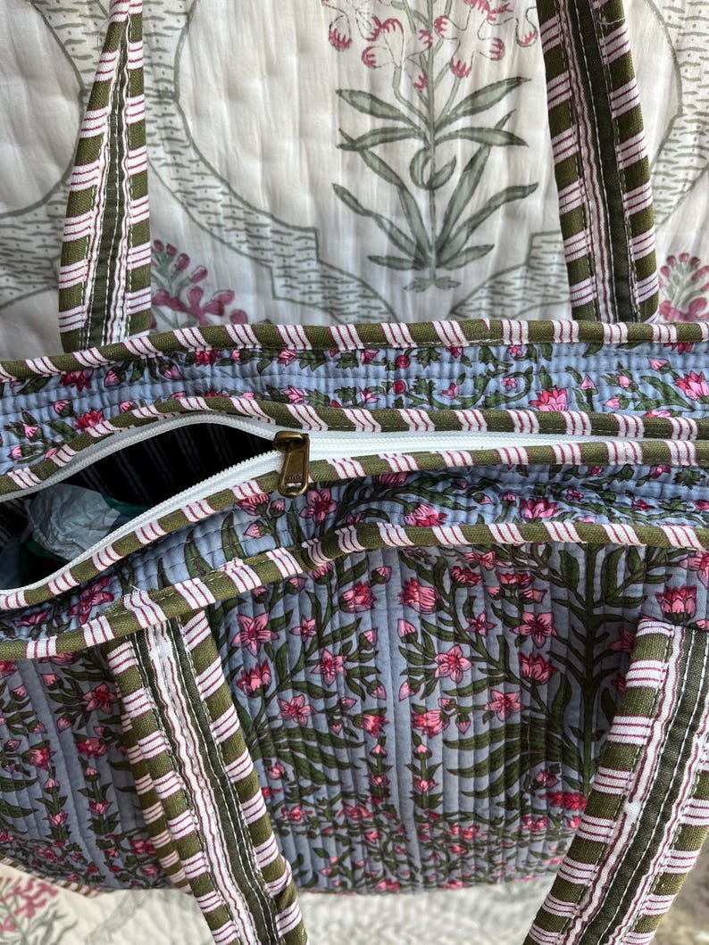 May include: A quilted tote bag with a floral pattern in pink and green on a light blue background. The bag has a white zipper and olive green straps with pink and white stripes. The bag is made of a soft, quilted material.