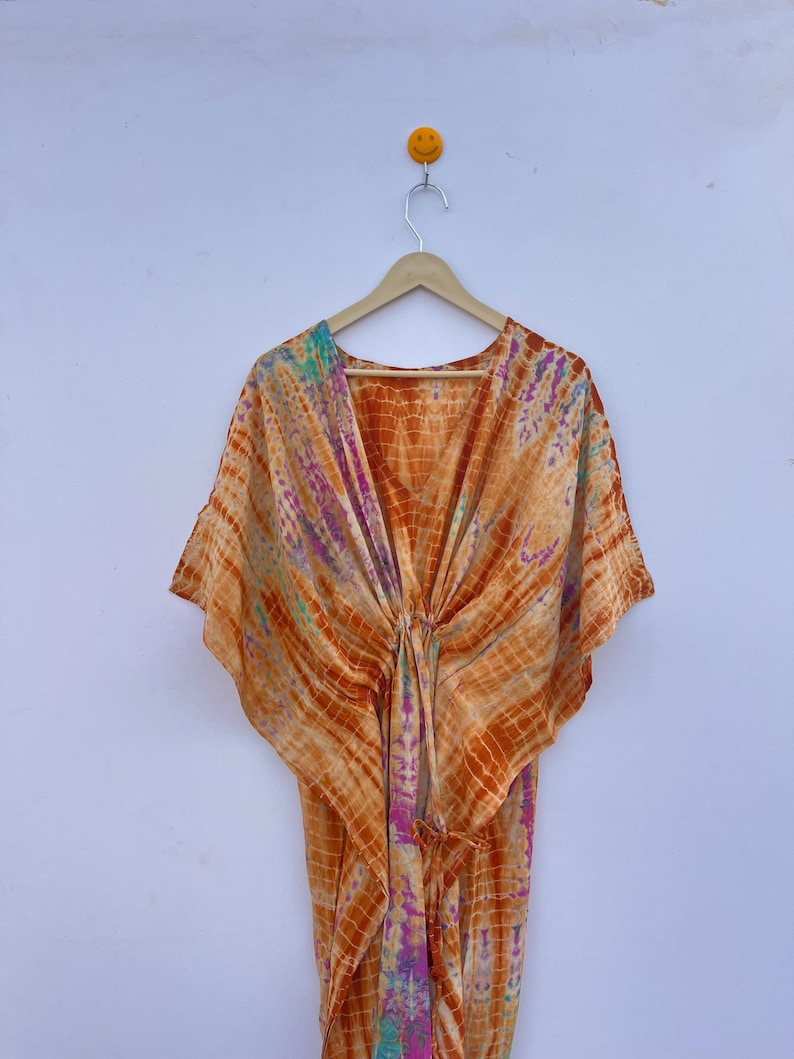 Puede incluir: Un vestido caft&aacute;n naranja te&ntilde;ido anudado colgado de una percha de madera. El vestido presenta un escote en V y mangas anchas. La tela tiene un patr&oacute;n de colores naranja, rosa, morado y turquesa. El vestido est&aacute; anudado a la cintura.