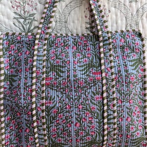 May include: A quilted tote bag with a floral pattern in pink and green on a light blue background. The bag has matching handles with a striped design in white, green, and pink. The bag is hanging on a black hanger.