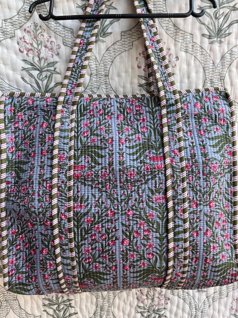 May include: A quilted tote bag with a floral pattern in pink and green on a light blue background. The bag has matching handles with a striped design in white, green, and pink. The bag is hanging on a black hanger.