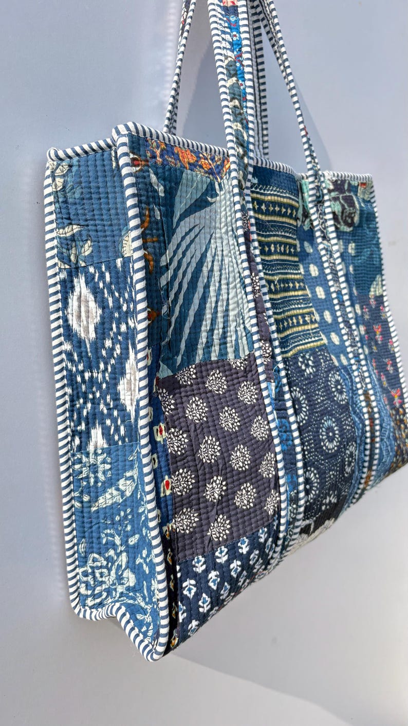 May include: A patchwork tote bag with a variety of blue fabric patterns. The bag features a quilted design with white and blue striped trim and handles. The bag is approximately 15 inches wide and 14 inches tall.