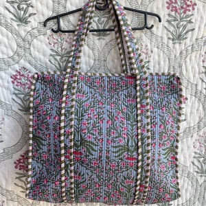 May include: A quilted tote bag with a floral pattern in shades of blue, pink, and green. The bag has a rectangular shape with two handles and a striped trim. The bag is hanging on a black hanger.