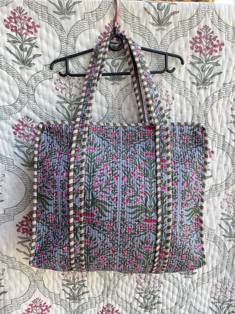 May include: A quilted tote bag with a floral pattern in shades of blue, pink, and green. The bag has a rectangular shape with two handles and a striped trim. The bag is hanging on a black hanger.