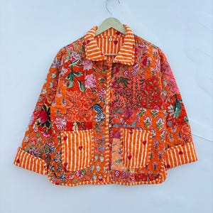 Puede incluir: Una chaqueta acolchada con un diseño de patchwork en naranja vibrante con varios patrones florales y botánicos. La prenda incluye un cuello y puños con rayas blancas y naranjas, dos bolsillos delanteros con pequeños detalles de corazones rojos y botones.