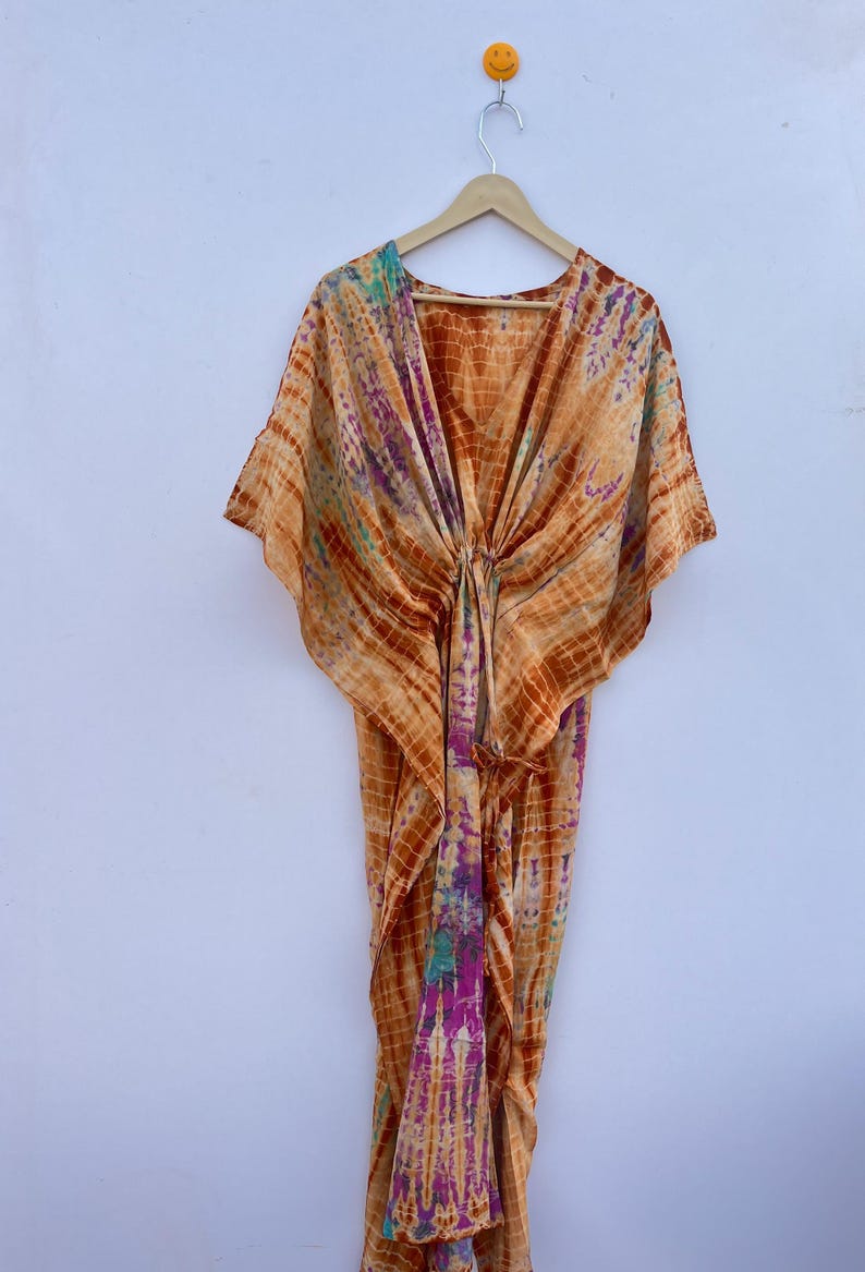 Puede incluir: Un caft&aacute;n te&ntilde;ido anudado en tonos naranja, morado y turquesa cuelga de una percha de madera. La prenda fluida presenta un cuello en V y mangas anchas. La tela est&aacute; estampada con l&iacute;neas horizontales y verticales, creando un efecto de cuadr&iacute;cula.