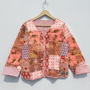 Peut inclure: Veste patchwork à col en V et manches longues. La veste présente divers motifs floraux et animaliers dans des tons de rose, d'orange et de jaune. Les poignets et la doublure intérieure ont un motif à rayures rouges et blanches.