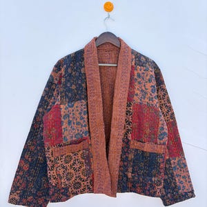 Könnte beinhalten: Eine langärmelige Jacke mit Patchwork-Design und verschiedenen Blumenmustern in Dunkelblau, Rot und Orange. Das Kleidungsstück hat einen breiten, einfarbigen orange-braunen Kragen, zwei Vordertaschen und sichtbare Steppstichdetails.