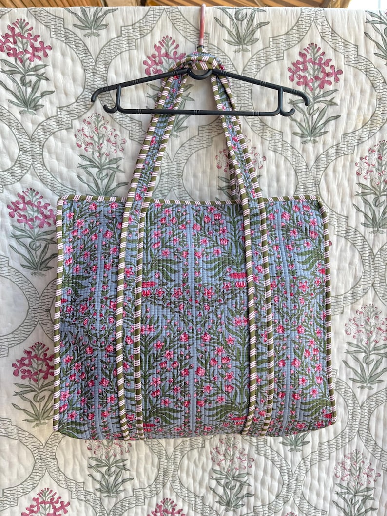 May include: A quilted tote bag with a floral pattern in pink and green on a light blue background. The bag has matching handles and trim. The bag is hanging on a black hanger against a floral patterned background.