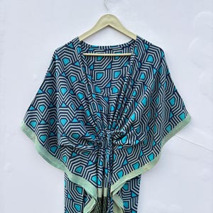 May include: A kaftan featuring a geometric pattern in shades of dark blue, light blue, and grey. The garment has a V-neckline, wide kimono-style sleeves, and a central tie detail at the waist that gathers the fabric, creating a draped silhouette.
