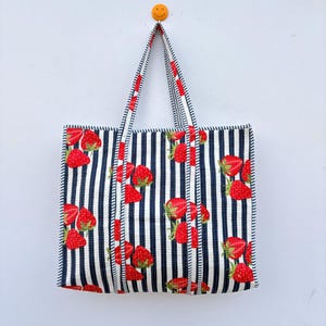 May include: A tote bag with a navy and white striped design, decorated with a bright red strawberry pattern. The bag has matching striped handles and is set against a white backdrop.