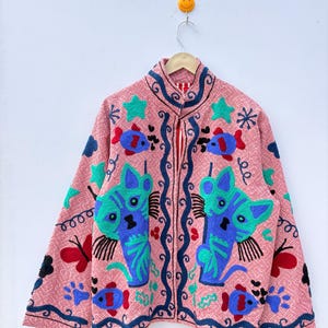 May include: Pink embroidered jacket featuring a repeating diamond pattern. The jacket has a high collar and a zipper closure. It is decorated with blue and green cat and fish designs, along with stars and butterflies.