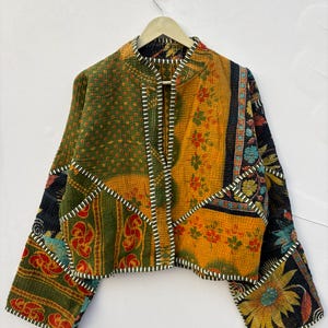 May include: A cropped jacket made from quilted fabric featuring a patchwork of green, yellow, and black panels with floral and geometric patterns. The garment has long sleeves, a mandarin collar, and white and black striped trim along the edges.
