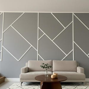 Könnte beinhalten: Ein modernes Wohnzimmer mit einer grauen Wand mit einem geometrischen weißen Zierleistendesign. Vor der Wand steht ein beiges Sofa, davor ein runder Holztisch und ein Teppich. Dekorative Gegenstände und Pflanzen stehen auf dem Tisch.