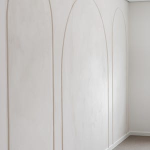 Könnte beinhalten: Eine weiße Wand mit drei bogenförmigen Dekorationselementen. Die Bögen sind mit einer dünnen, hellfarbigen Leiste umrandet. Die Wand hat eine strukturierte Oberfläche, und der Raum hat eine neutrale Farbpalette.