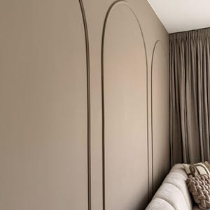 Könnte beinhalten: Innenraum mit einer Wand mit drei bogenförmigen dekorativen Paneelen in einem neutralen Beige. Die Wand grenzt an ein helles Sofa und braune Vorhänge, wodurch ein stilvolles und kohärentes Design entsteht.