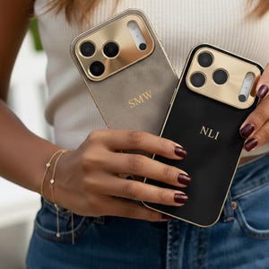 Puede incluir: Dos fundas de teléfono personalizadas. Una es gris con detalles dorados y las iniciales "SMW". La otra es negra con detalles dorados y las iniciales "NLI". Ambas fundas tienen un marco dorado para la cámara.