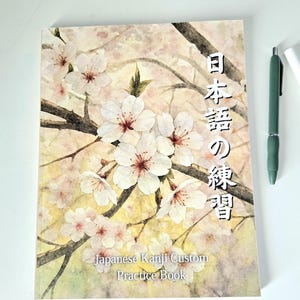 May include: A Japanese Kanji Custom Practice Book with a watercolor illustration of white cherry blossoms on a light background. The book has Japanese text on the cover. A green pen and white marker are on the right.