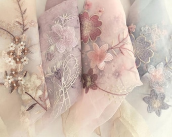 May include: Four rolls of sheer fabric in pastel colors, each embroidered with floral designs. The colors include cream, lavender, pink, and gray. The floral embroidery features delicate details and varying shades of pink, purple, and cream.
