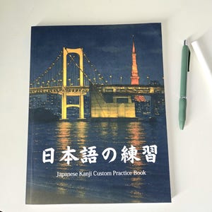 May include: A Japanese Kanji Custom Practice Book with a night scene of a bridge and tower. The book cover is dark blue with white Japanese characters and English text. A green pen rests on the white surface.