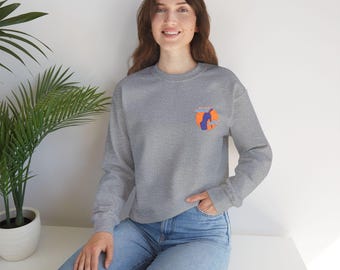 May include: A heather gray sweatshirt with a small graphic on the upper left chest. The graphic features a blue silhouette inside an orange circle with text. The model is wearing blue jeans and is sitting on a white surface.