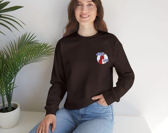 May include: A dark brown sweatshirt with a small embroidered patch on the upper left chest. The patch features a red silhouette of a dancer inside a white circle with blue text that reads "exotic dancer" and "since 2009". The model is wearing blue jeans.