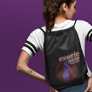 Può includere: Zaino nero con coulisse con la scritta "exotic pole dancer" e un disegno di scarpa con tacco alto viola. La persona indossa una maglietta bianca con strisce nere sulle maniche.