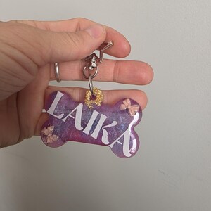Puede incluir: Etiqueta para mascotas en forma de hueso con el nombre "LAIKA" en letras blancas. La etiqueta tiene un diseño veteado morado y azul con pequeños lazos rosas y un acento dorado brillante. Está sujeta a un cierre plateado.