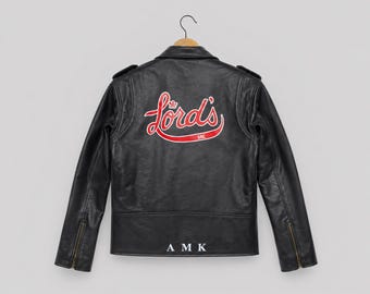 May include: A black leather jacket featuring a red and white script logo on the back that reads Lord's with a small crown icon above the L and the letters SAC below. The jacket includes shoulder epaulets, zippered cuffs, and the letters A M K in white.