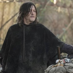 May include: A person wearing a black hooded poncho stands next to a motorcycle. The poncho has a textured appearance. The motorcycle has a round mirror and a bundle of supplies attached to the front.