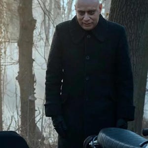 May include: A man wearing a long black coat and black gloves stands outdoors. The coat has a button closure and a collar. The man has a shaved head and is looking down. A motorcycle seat is visible in the foreground.