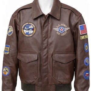 Könnte beinhalten: Braune Leder-Bomberjacke mit mehreren Aufnähern, darunter "Tuskegee Airmen" und "U.S. Army Air Corps". Sie hat zwei Vordertaschen, einen gerippten Saum und Bündchen. Die Jacke hat einen klassischen Kragen und einen Reißverschluss. Ein amerikanischer Flaggenaufnäher ist sichtbar.