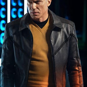 May include: A black leather jacket with a V-neck yellow shirt underneath. The jacket has a textured shoulder detail and a contrasting design on the sleeves. The man in the image has gray hair and is looking to the left.