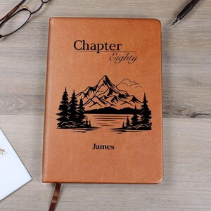 Puede incluir: Un diario de cuero marrón con las palabras "Chapter Eighty" y el nombre "James" impresos en la portada. Una escena de montaña y lago en negro también está impresa en la portada. Una cinta de marcador marrón es visible.