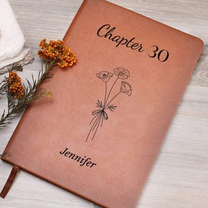 May include: A brown leather journal with the text "Chapter 30" and the name "Jennifer" in black script. A floral illustration is below the text. A brown ribbon bookmark is visible.