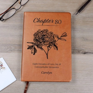 Puede incluir: Un diario de cuero marrón con la inscripción "Chapter 80" y un diseño floral. El diario también incluye el texto "Eight Decades of Love, Joy & Unforgettable Memories" y el nombre "Carolyn".