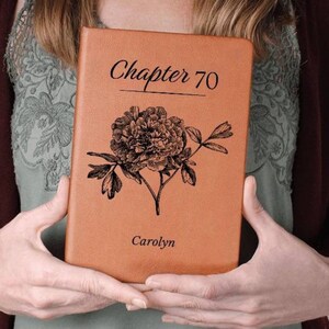 May include: A tan-colored book with the words "Chapter 70" and "Carolyn" printed on the cover. A detailed black floral illustration is centered on the cover. The book is held by a person.