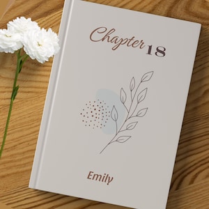 Pode incluir: Um livro de capa dura com uma capa cinza claro. A capa apresenta o texto "Chapter 18" em marrom, uma ilustração floral e o nome "Emily". Flores brancas no fundo.