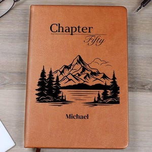 Puede incluir: Un diario de cuero marrón con las palabras "Chapter Fifty" y "Michael" impresas en la portada. Una escena de montaña y lago negra está grabada debajo del texto. El diario está cerrado y tiene una cinta de marcador.