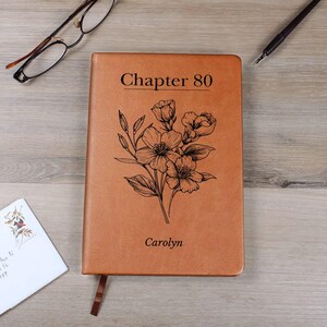 Puede incluir: Un diario de cuero marrón con el texto "Chapter 80" y el nombre "Carolyn" grabado en la portada. Una ilustración floral también está grabada en la portada. Una cinta de marcador marrón es visible.