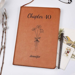 May include: A brown leather journal with the text "Chapter 40" and the name "Jennifer" engraved on the cover. A floral illustration is also featured. A black pen rests beside the journal.