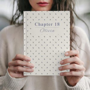Peut inclure: Un livre à couverture rigide crème avec un motif répété de petites fleurs bleues. Le titre, "Chapter 18 Olivia", est imprimé en bleu clair. Le livre est tenu par une personne.