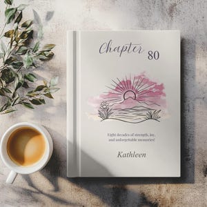 Puede incluir: Un libro de tapa dura con el título "Chapter 80" y una ilustración de acuarela de un amanecer sobre un paisaje. La portada del libro también incluye el texto "Eight decades of strength, joy, and unforgettable memories!" y el nombre "Kathleen".