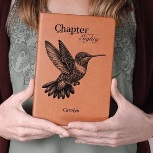 Puede incluir: Un libro de color marrón claro con las palabras "Chapter Eighty" y el nombre "Carolyn" impresos. Una ilustración negra detallada de un colibrí aparece en la portada. El libro está sostenido por una persona.